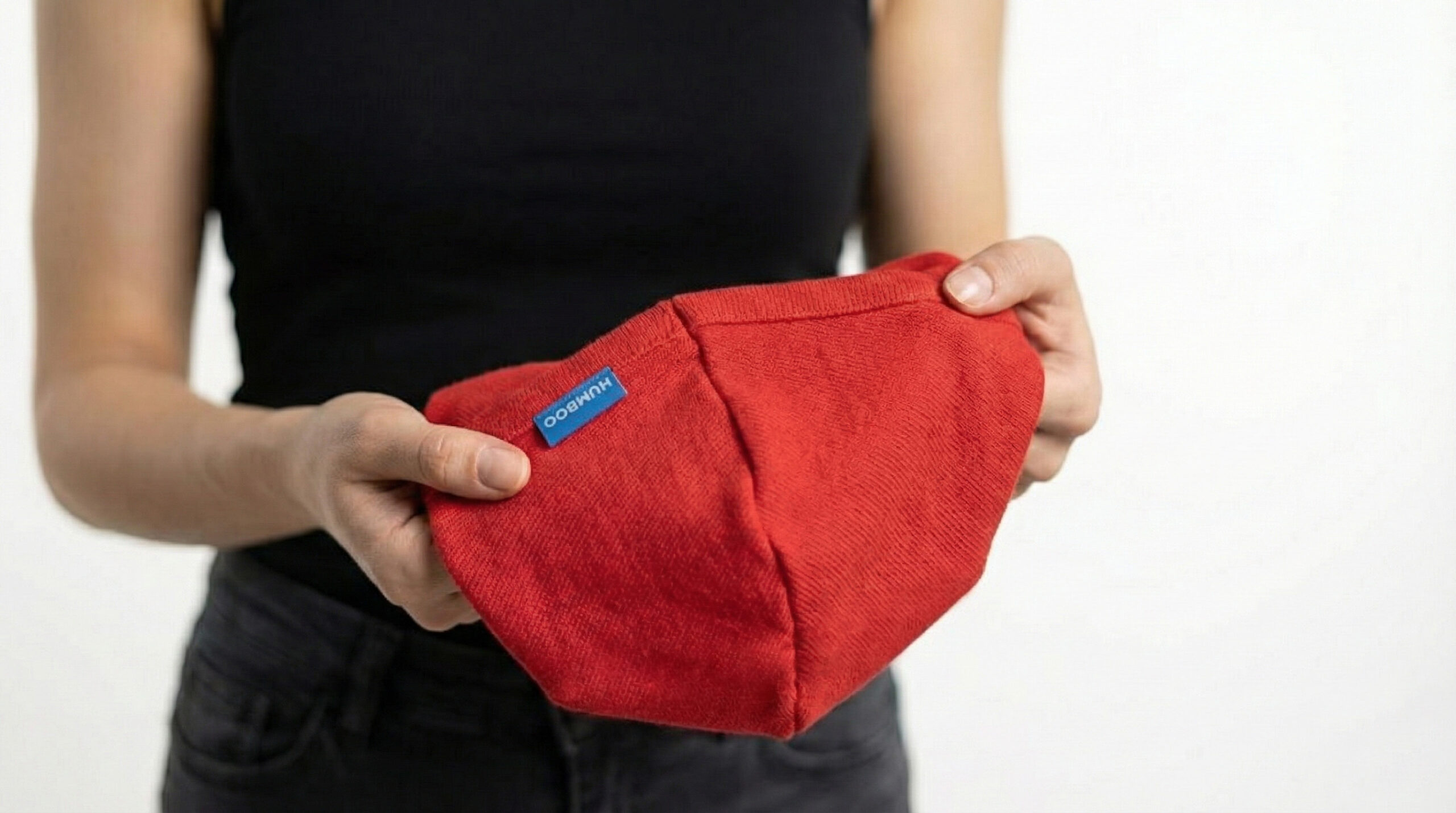 La foto muestra a una persona sosteniendo en sus manos un pasamontañas rojo de algodón, en cuyo lateral, en el lugar habitual del cuello, hay una etiqueta rectangular azul característica con la inscripción blanca «HUMBOO»; el producto se presenta sobre un fondo blanco neutro, lo que permite observar con detalle la textura del tejido y la ubicación de la marca, típica de los modelos de algodón como OptiBalance o FlexyPrime.