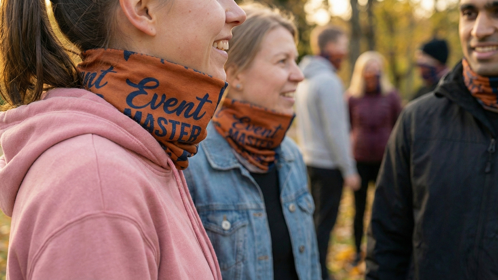 Los participantes sonrientes de un evento al aire libre en un parque otoñal, llevando idénticos pasamontañas multifuncionales naranjas y azules con el estampado personalizado «Event Master».