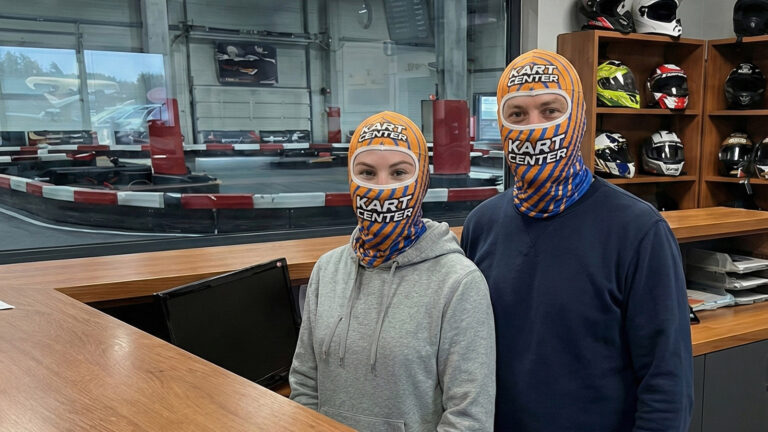 Para w recepcji toru kartingowego KART CENTER ubrana w termoaktywne kominiarki z logo -poliestrowe z pomarańczowo-niebieskim nadrukiem sublimacyjnym jako alternatywę dla modeli bawełnianych. W tle widoczna infrastruktura toru i kaski.