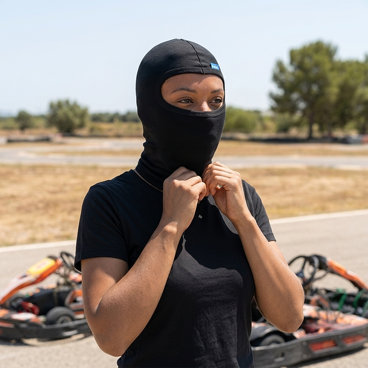 Kobieta w czarnej bawełnianej kominiarce Humboo stoi przy torze kartingowym przed sesją, w tle rozmyty gokart i słoneczna łaka.