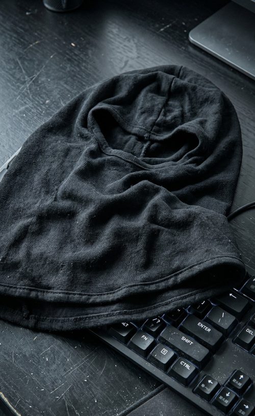 Zbliżenie na czarną, dzianinową maskę kominiarkę typu balaclava, spoczywającą na porysowanym, czarnym biurku gamingowym. Obok maski widoczna jest krawędź podświetlanej klawiatury mechanicznej, podkreślając taktyczny i gamingowy charakter scenerii.